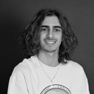 Jeune homme souriant avec de longs cheveux bruns, portant un t-shirt blanc et un collier.