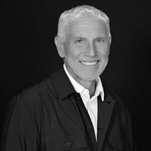 Portrait en noir et blanc d'un homme mûr aux cheveux gris souriant, vêtu d'une veste sombre sur une chemise blanche.