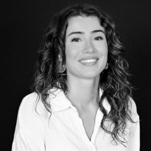 Portrait en noir et blanc d'une femme souriante aux cheveux bouclés portant une chemise blanche sur fond noir.