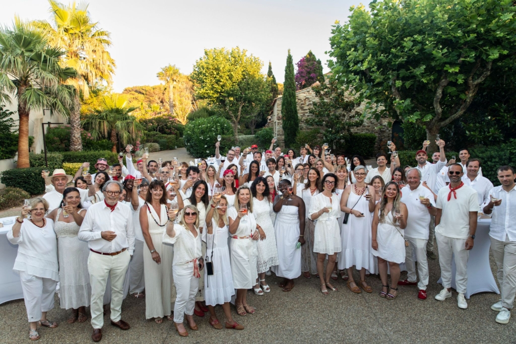 Un grand groupe de personnes en tenues blanches lève des verres à l'extérieur lors d'une fête ensoleillée.
